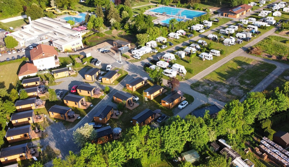 An aerial view captures a well-organized camping park, featuring numerous caravan sites alongside cabins. The outdoor swimming pool is prominent, bordered by green areas. Surrounding trees provide a natural setting, and pathways connect various sections of the park, indicating an inviting and spacious environment.
