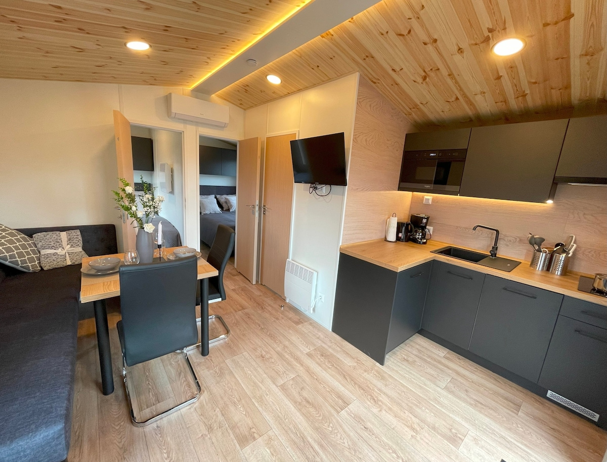 A modern interior features a cozy seating area with a dark sofa and a dining table set for two. The kitchen is equipped with sleek black cabinets, an electric stove, and essential appliances. Warm wood accents highlight the ceiling and floor, creating an inviting ambiance.