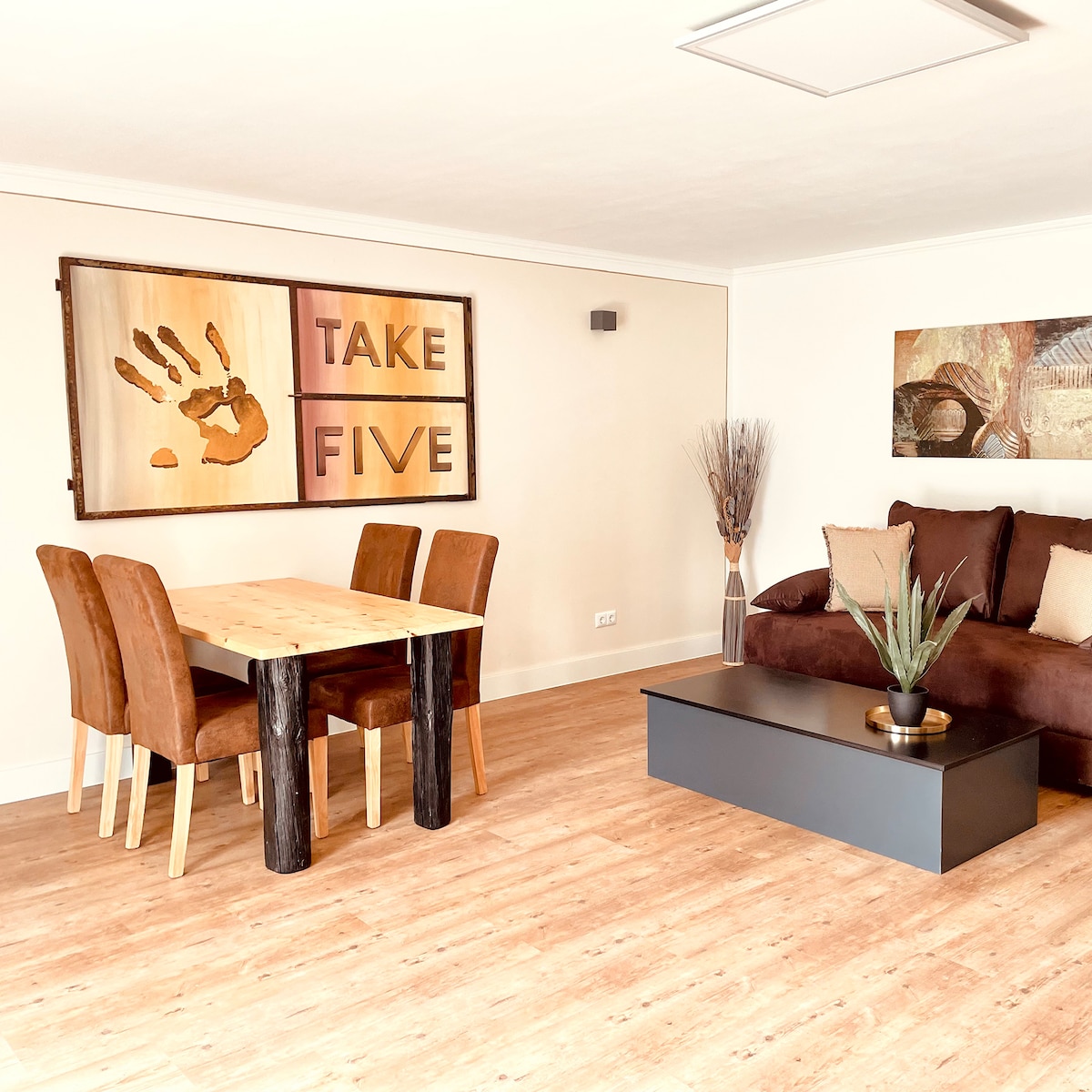 A dining area features a wooden table surrounded by four brown chairs. A comfortable brown sofa is positioned nearby, complemented by a coffee table with a small plant. The wall art displays the phrase 'TAKE FIVE', adding a distinct touch to the space.