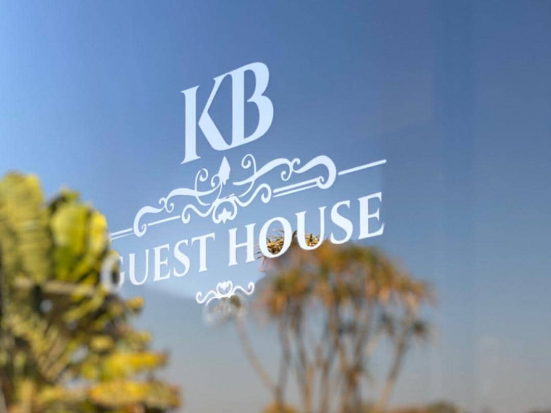 The exterior features a clear reflection of palm trees against a blue sky, with a decorative sign displaying 'KB GUEST HOUSE' prominently featured. The sign's elegant design adds a welcoming touch to the entrance.