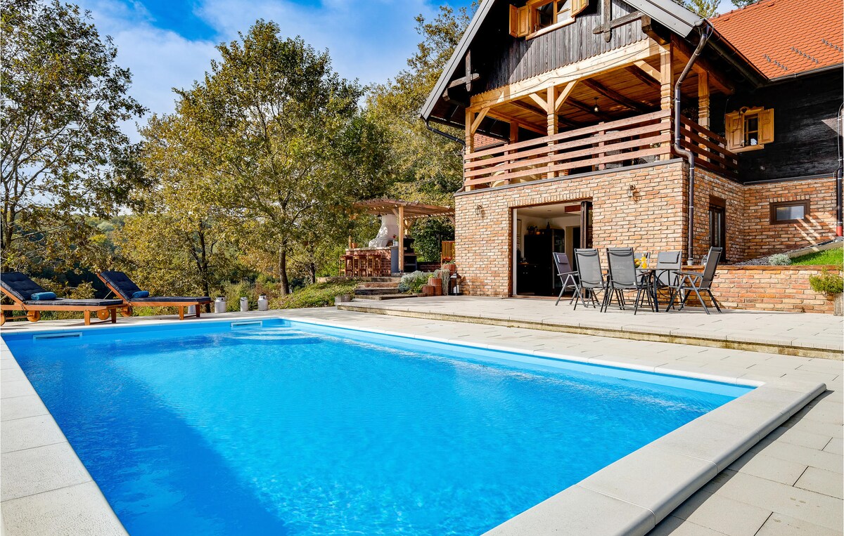 A private outdoor swimming pool is framed by a spacious terrace, featuring several lounge chairs. The vacation home displays a modern rustic style with a wooden balcony and brick exterior. Surrounding trees offer a natural setting, enhancing the outdoor relaxation area.