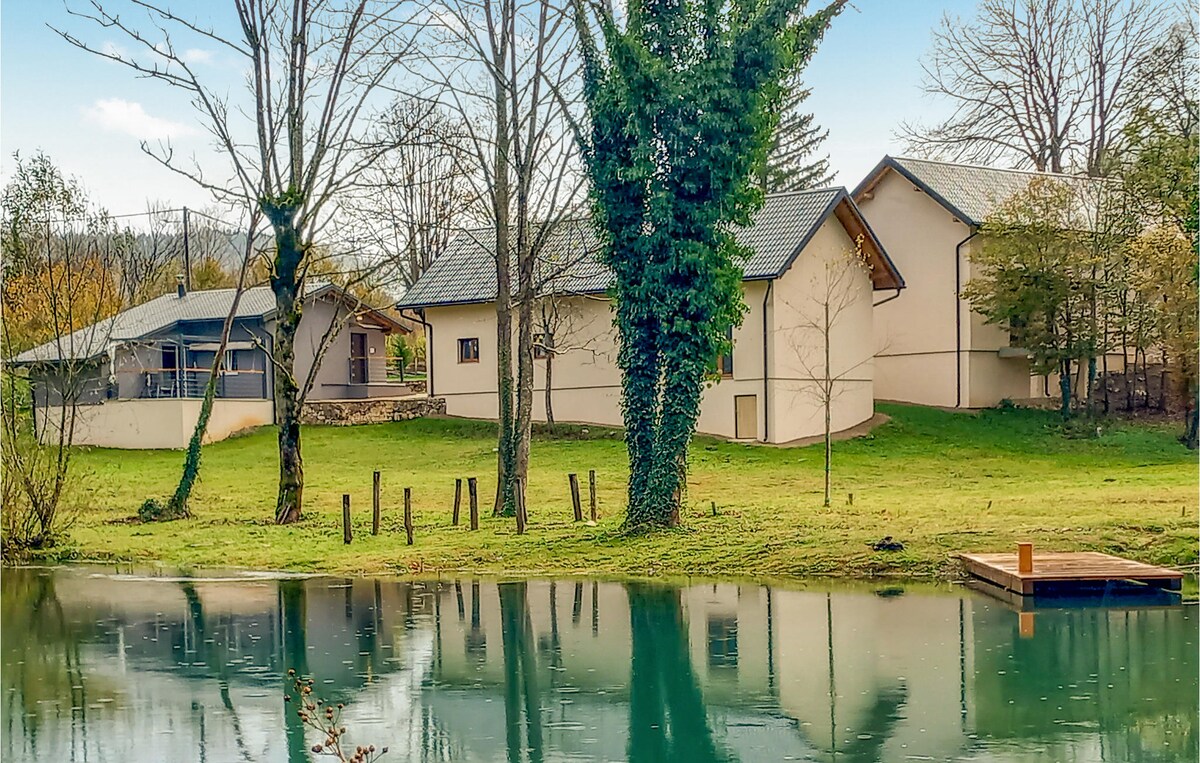 A modern house is situated next to a calm body of water, surrounded by trees. The building features a light exterior with a sloped roof. A wooden dock extends into the water, reflecting the serene landscape. The greenery enhances the peaceful atmosphere.