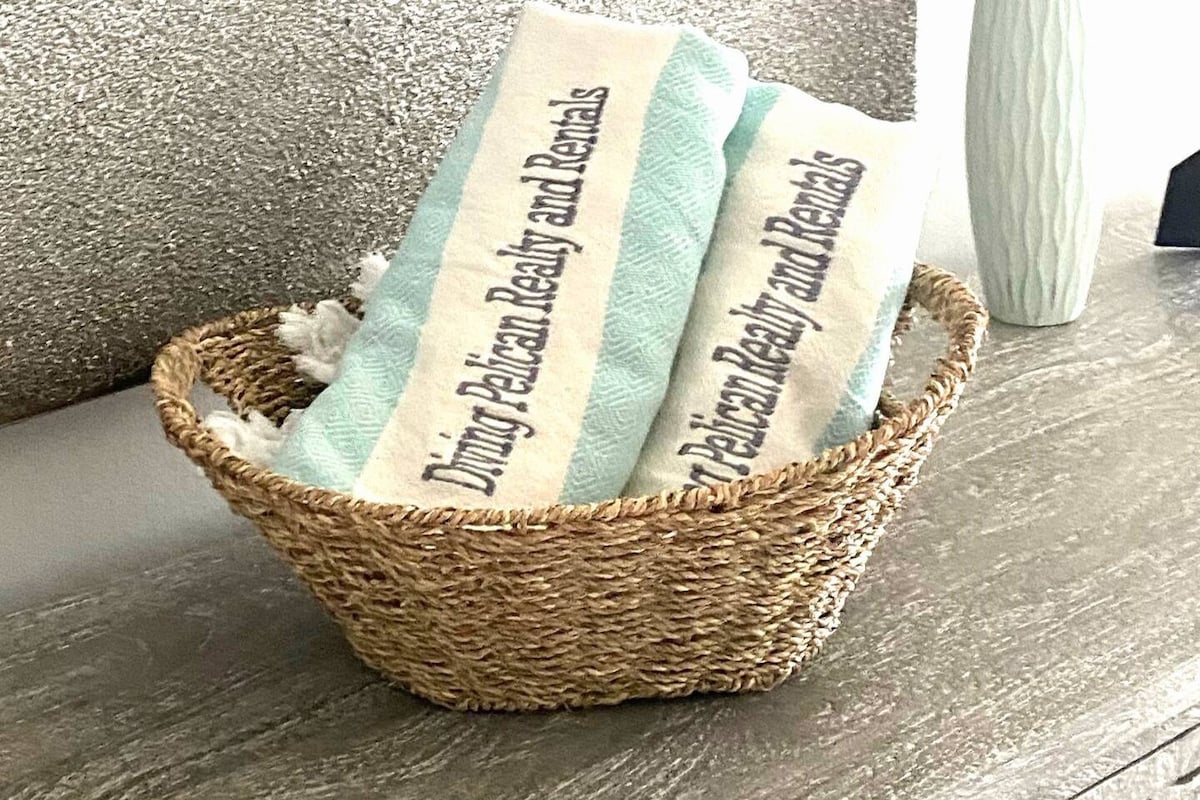 A woven basket is filled with neatly arranged towels, displaying the text 'Diving Pelican Realty and Rentals.' The towels are presented with a soft blue and white color scheme, complementing the neutral tones of the surrounding decor.