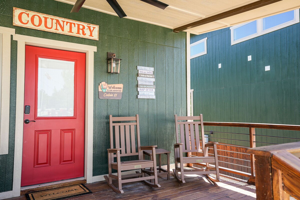 The cabin entrance features a vibrant red door and a welcoming sign. Two rocking chairs are placed on the porch, offering a cozy spot to relax. The green wooden exterior and decorative elements create an inviting aesthetic.