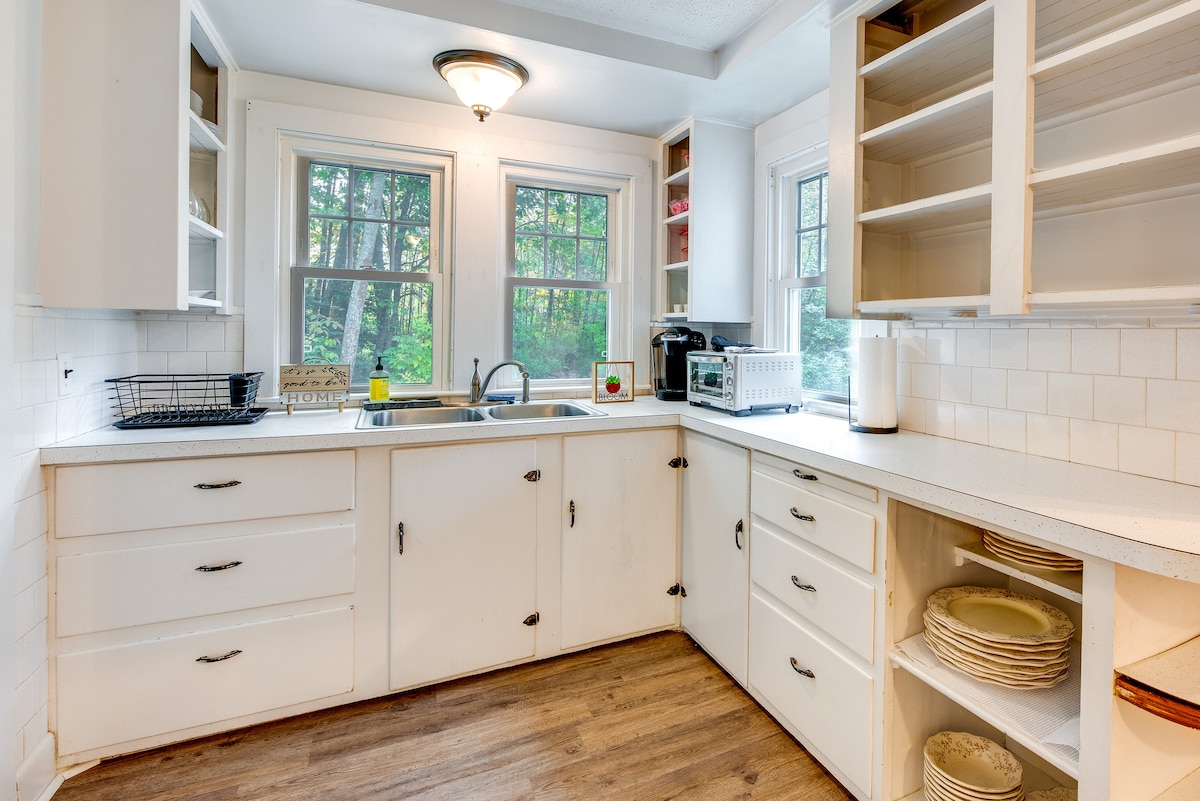 The kitchen features white cabinetry and countertops, providing ample storage and workspace. A double sink is situated under two windows that allow natural light to illuminate the area. Appliances such as a microwave and a coffee maker are visible, along with dishware neatly arranged on open shelves.