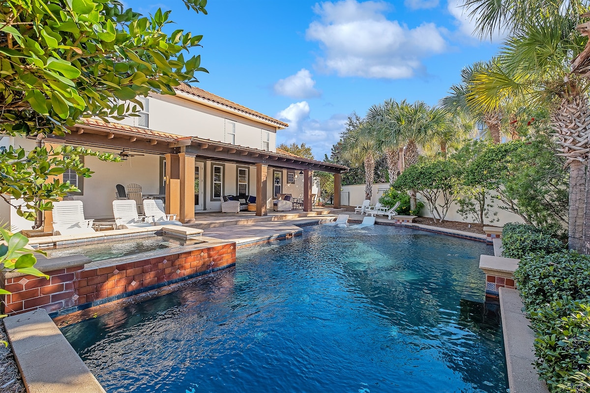 The tranquil pool area features a spacious layout surrounded by palm trees and lush landscaping. A hot tub is situated nearby, and patio loungers are arranged along the edge. The home is visible in the background, showcasing its elegant architecture under a clear blue sky.