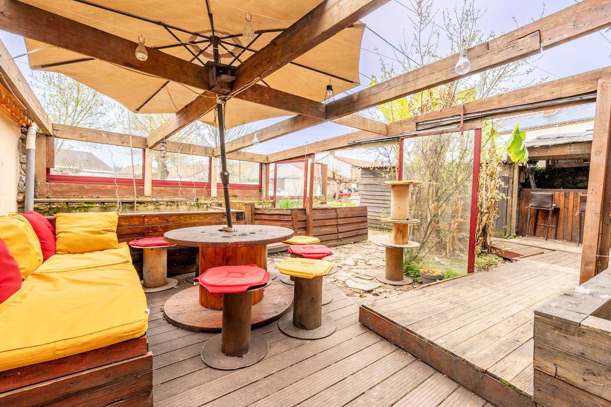 An outdoor seating area is showcased, featuring a round table surrounded by colorful stools. A large shaded area is provided by a canopy overhead. Wooden pathways lead to various sections, including a small garden with plants and a cat tree.