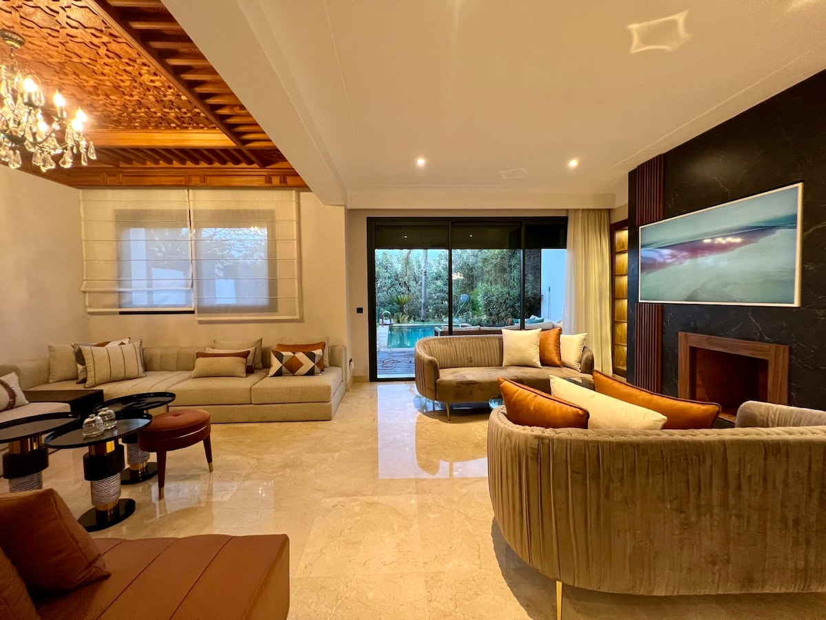 A spacious living room features luxurious seating arrangements with plush sofas and chairs. Natural light filters through large glass doors that provide a view of the outdoor area. Elegant decor elements include a decorative ceiling and a modern fireplace.
