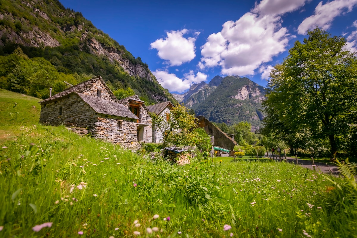 A rustic stone chalet is surrounded by lush greenery and towering mountains. The structure features a traditional stone facade with a slate roof, blending harmoniously with the natural landscape. Bright clouds drift in the blue sky above, enhancing the serene feeling of the outdoor space.