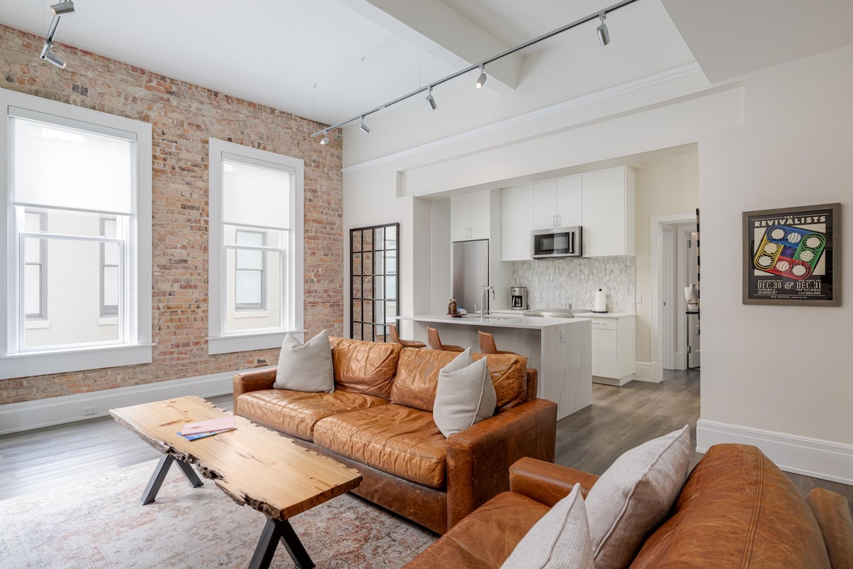 A spacious living area features a mix of leather sofas and a wooden coffee table, complemented by exposed brick walls. Large windows allow natural light to fill the space. An open kitchen is visible in the background, showcasing modern appliances and white cabinetry.