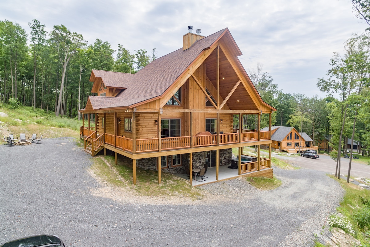 The exterior of Hilltop Cottage is shown, featuring a spacious wooden structure with multiple gables and expansive decks. Surrounded by trees, the wrap-around deck is visible, designed for relaxation. A gravel driveway and nearby homes hint at the community setting.