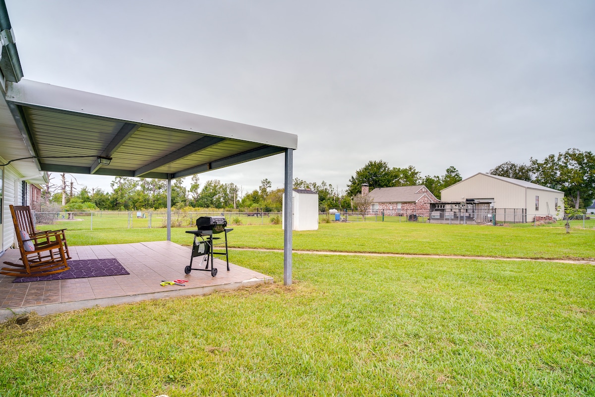 A covered patio features a grill and two rocking chairs, providing an outdoor space to relax. The expansive yard is visible, showcasing a grassy area, with nearby structures and trees in the background. The setting offers a peaceful retreat in the open air.