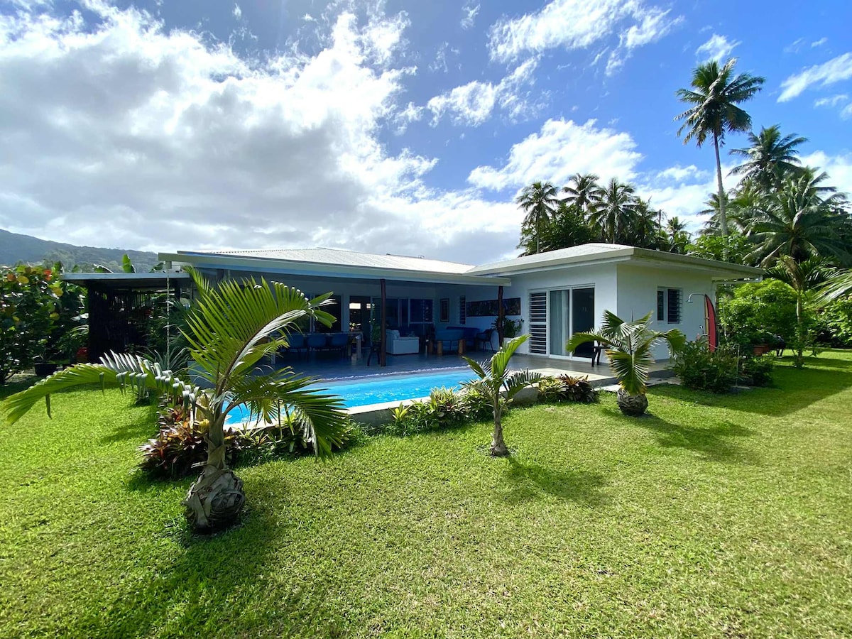 The exterior of a house is depicted, featuring a large, fenced garden with tropical plants and a swimming pool. A covered terrace can be seen, offering views of the pool and garden. Lush greenery and palm trees surround the property, under a partly cloudy sky.