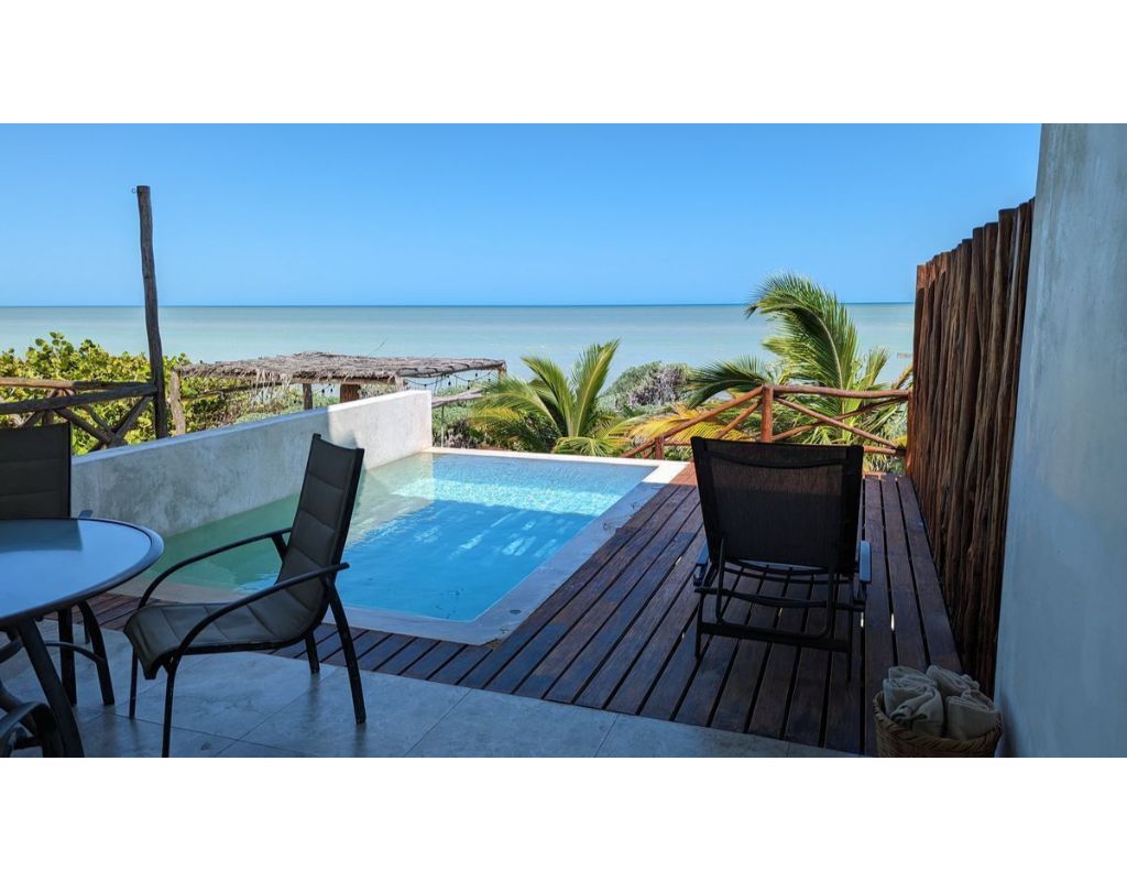 An outdoor terrace features a private pool surrounded by wooden decking, complemented by tropical greenery. A round dining table and a lounge chair are positioned for relaxation, with the serene ocean visible in the background under a clear blue sky.