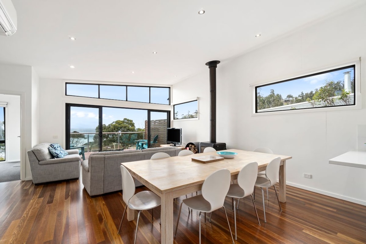 An open-plan living and dining area features a large wooden table surrounded by white chairs. A comfortable grey sofa is positioned near a wood fire stove. Floor-to-ceiling windows allow natural light to fill the space, offering views of the surrounding landscape and ocean.