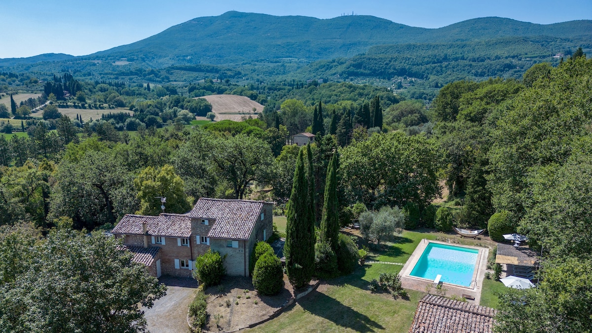 Aerial view of Casale Le Querce di Cetona showcases the rustic farmhouse with a terracotta roof amidst lush greenery. The property features a rectangular swimming pool surrounded by sun loungers and trees, inviting moments of relaxation in the serene Tuscan landscape.