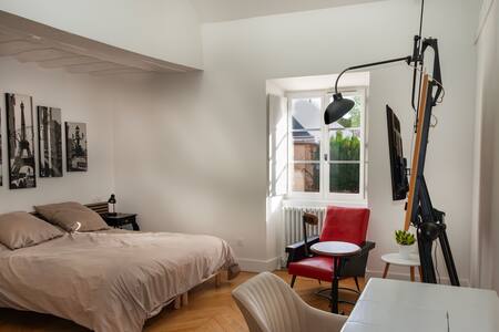 A spacious room features a large bed with neutral bedding, complemented by a bedside lamp and a small table. A modern chair in red adds a pop of color. Natural light filters through the window, which faces a tranquil outdoor view.
