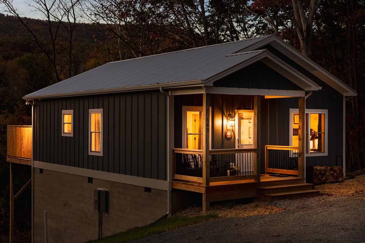 An inviting cabin exterior is illuminated in warm tones, showcasing a porch with steps leading up. Large windows offer a glimpse into the cozy interior. The structure is nestled among wooded surroundings, emphasizing a peaceful retreat in nature.