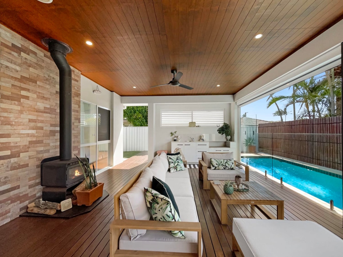An outdoor alfresco area features a wooden deck with comfortable seating arrangements and decorative cushions. A wood-burning stove is positioned against a stone feature wall. A lap pool is visible in the background, surrounded by palm trees and natural light.