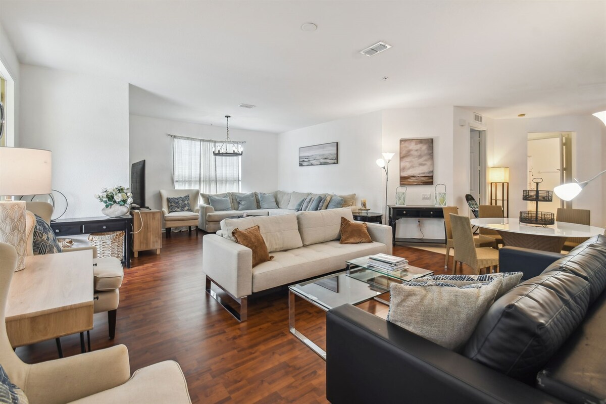 An open living area features a combination of seating options, including light-colored sofas and dark leather chairs. Natural light filters through a nearby window. A glass coffee table is positioned at the center, adding to the spaciousness of the room, which includes artwork on the walls.