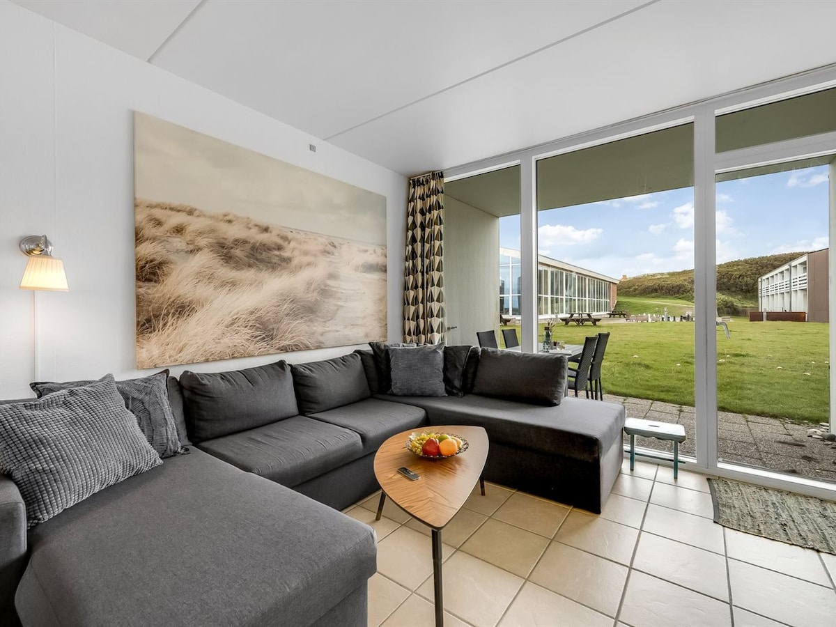 A spacious living room features a large sectional sofa complemented by a sleek coffee table. Natural light fills the area through expansive glass doors, offering a view of the outdoor space. A neutral-toned artwork adorns the wall, adding a calming touch.