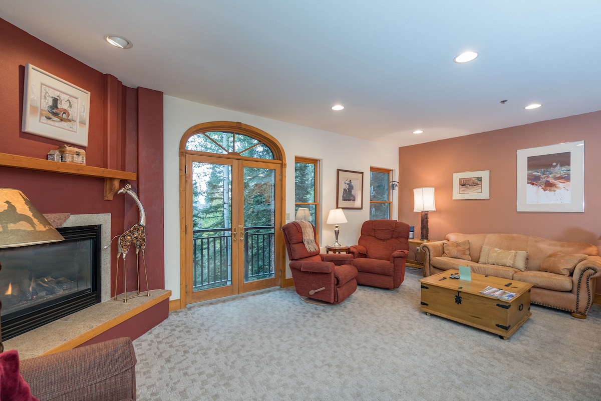 A living room features two reclining chairs and a plush sofa arranged around a wooden coffee table. A gas fireplace is positioned against a red accent wall. Large windows provide a forest view, with soft lighting from a nearby lamp creating a comfortable environment.