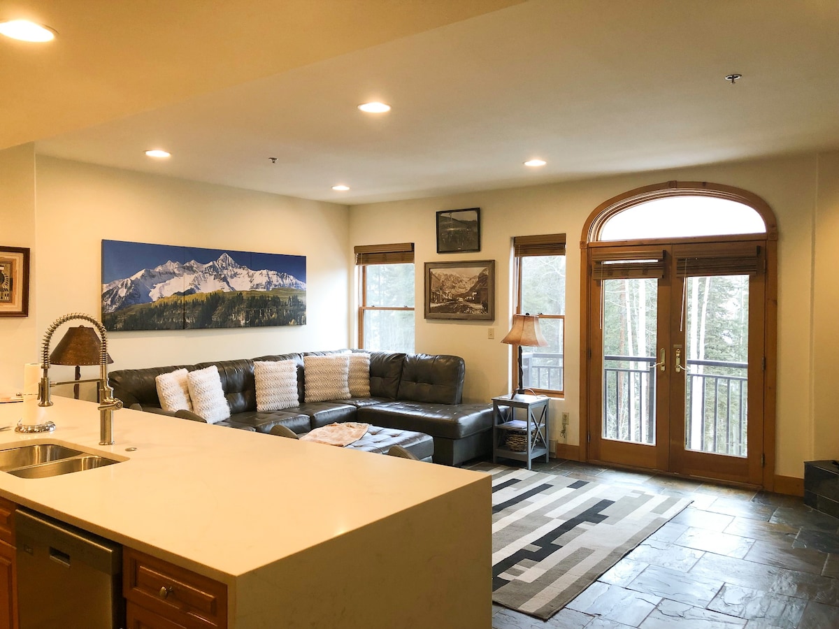 A spacious living area is visible, featuring a comfortable sectional couch and large windows with a view of the outdoors. A coffee table rests on a patterned rug, while double doors provide access to the private outdoor deck. The kitchen area includes a modern countertop.