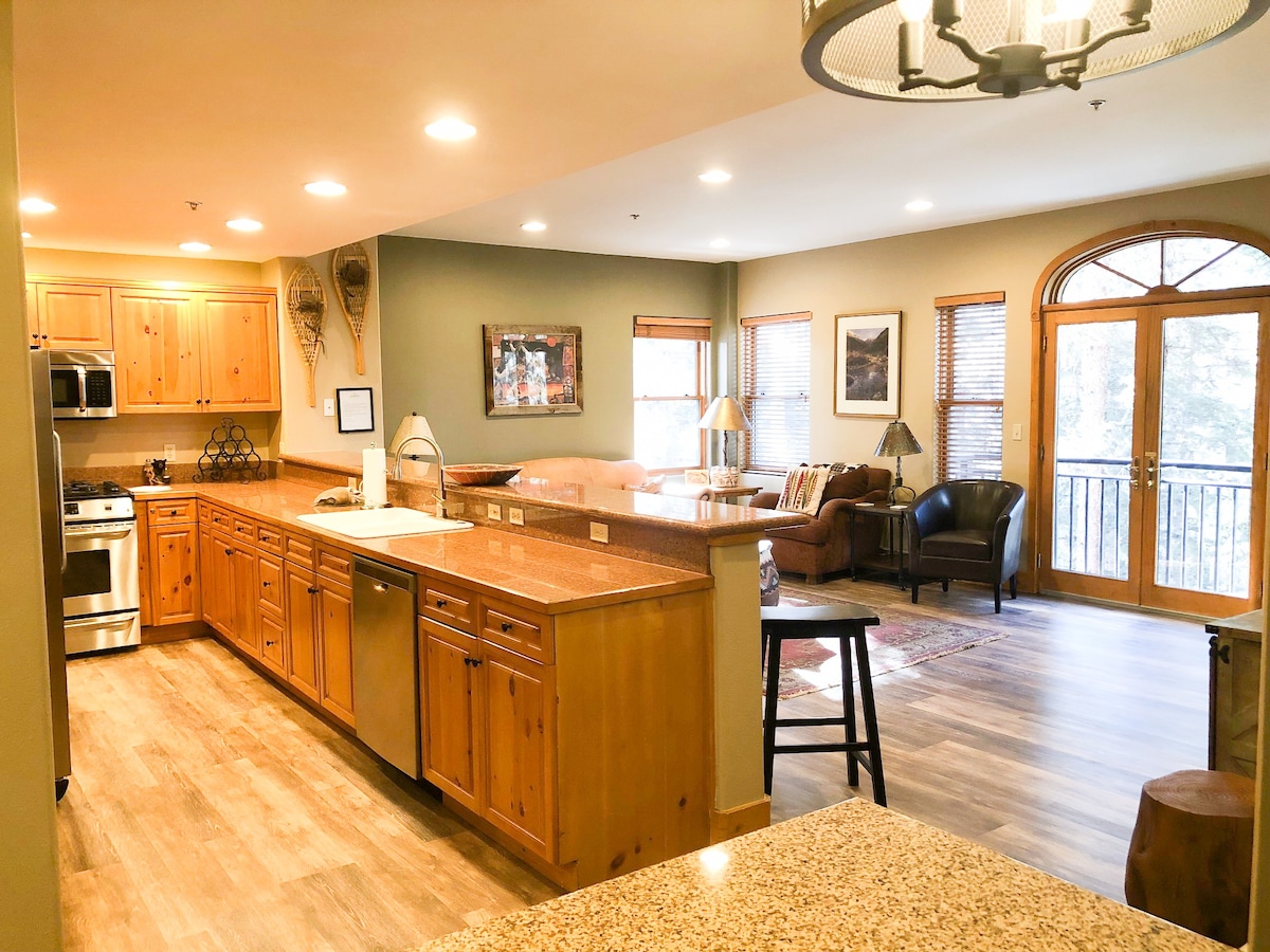 An open kitchen features wood cabinetry and granite countertops, with appliances including a refrigerator and stove. The dining area flows into a living space containing a seating area with a lamp and large windows offering a view of the balcony. Natural light highlights the warm tones of the wood.