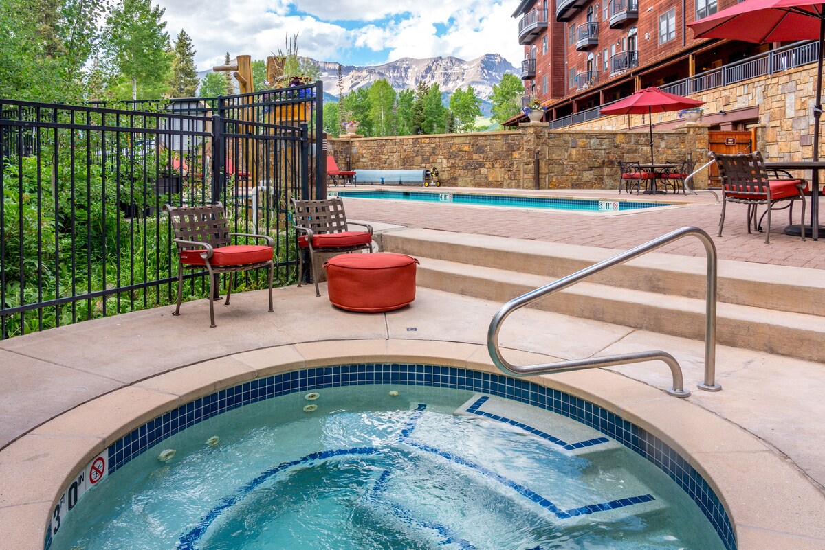 A spacious hot tub with bubbling water is surrounded by stonework and overlooks the outdoor heated pool. Red cushions are placed on nearby chairs, and shaded seating areas appear throughout the space. View of mountains can be seen in the background under a partly cloudy sky.