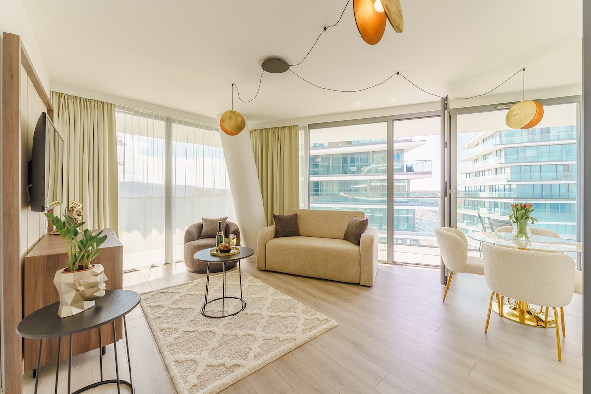 A spacious living area features large windows allowing natural light to fill the space. A comfortable sofa and armchair are positioned around a round coffee table, accompanied by a dining table set for four. Soft curtains drape elegantly beside the glass door leading to a balcony.