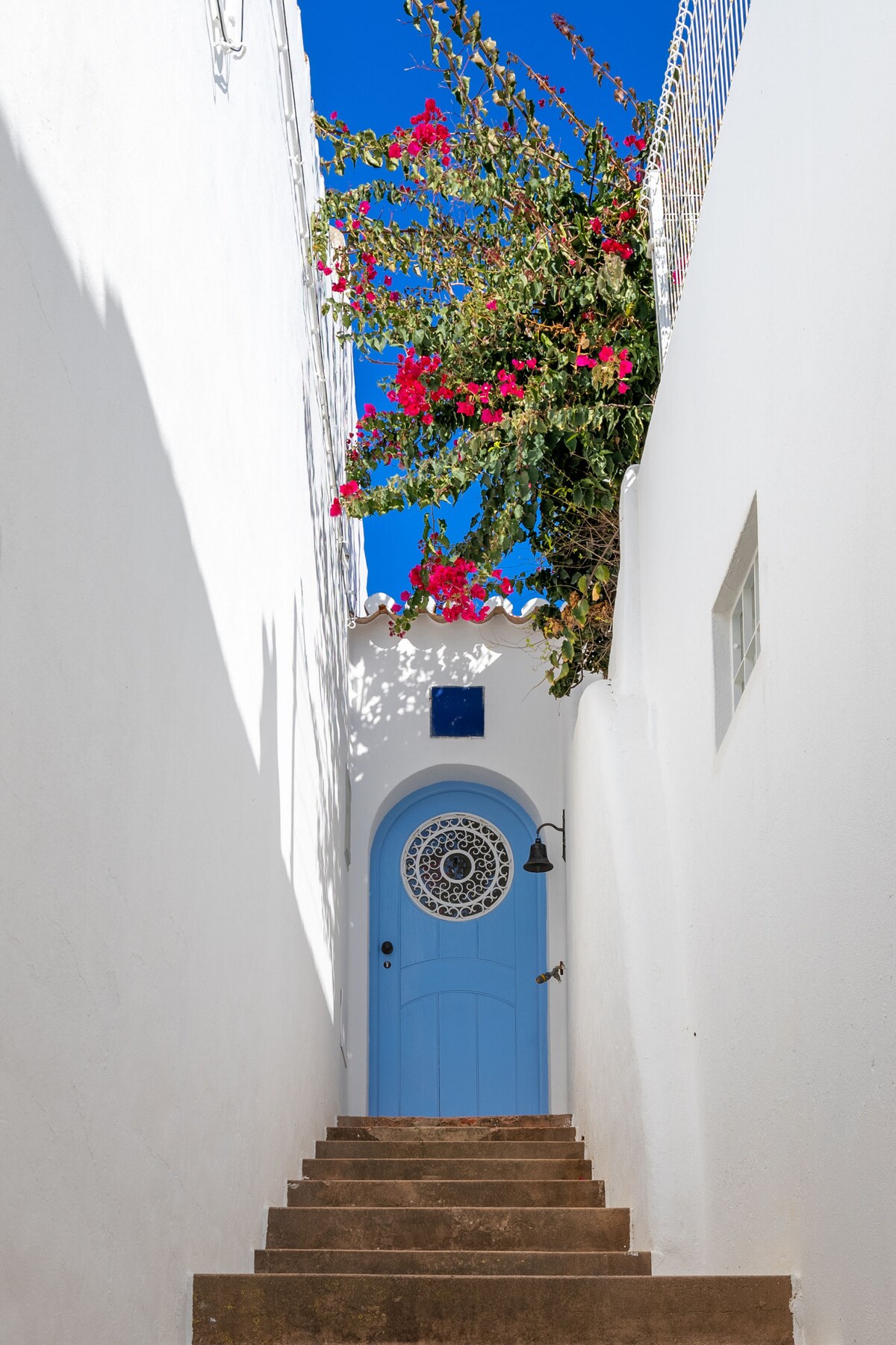 A narrow staircase is flanked by white walls, leading to a charming light blue door with a decorative circular window. Vibrant pink flowers cascade from above, contrasting against the clear blue sky.