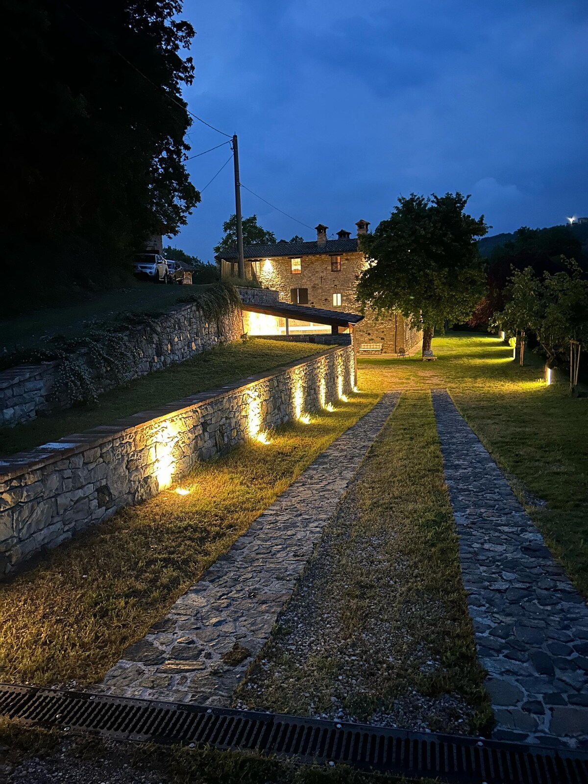 The exterior of the property is illuminated at night, highlighting a stone pathway lined with grass. The building is constructed from rustic materials, featuring multiple stories and a covered terrace. Ambient lighting enhances the landscaping along the pathway.