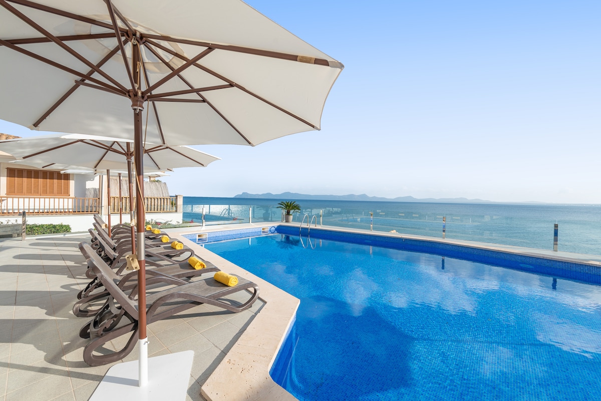 A spacious outdoor terrace features a large swimming pool with crystal clear water, surrounded by lounge chairs and umbrellas. The seafront offers expansive views of the calm ocean and distant islands, enhancing the serene atmosphere of the space.