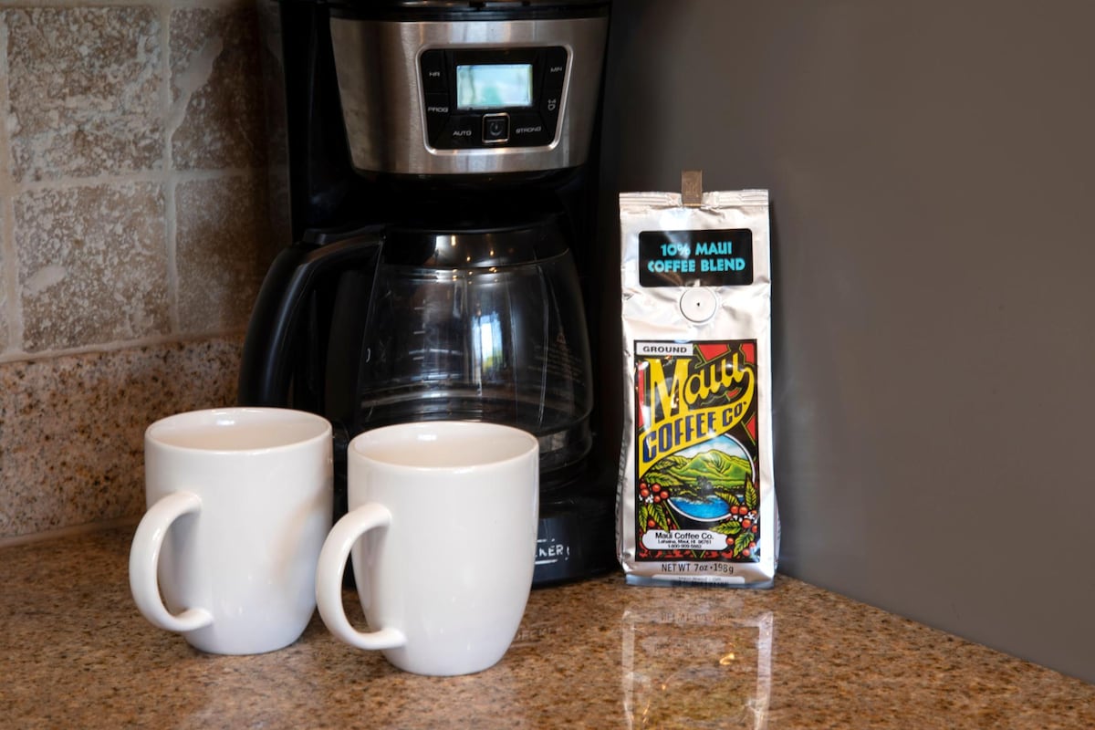 A coffee maker sits on a granite countertop next to two white mugs. Nearby, a bag of Maui coffee blend is displayed, highlighting the charming kitchen essentials available for guests.