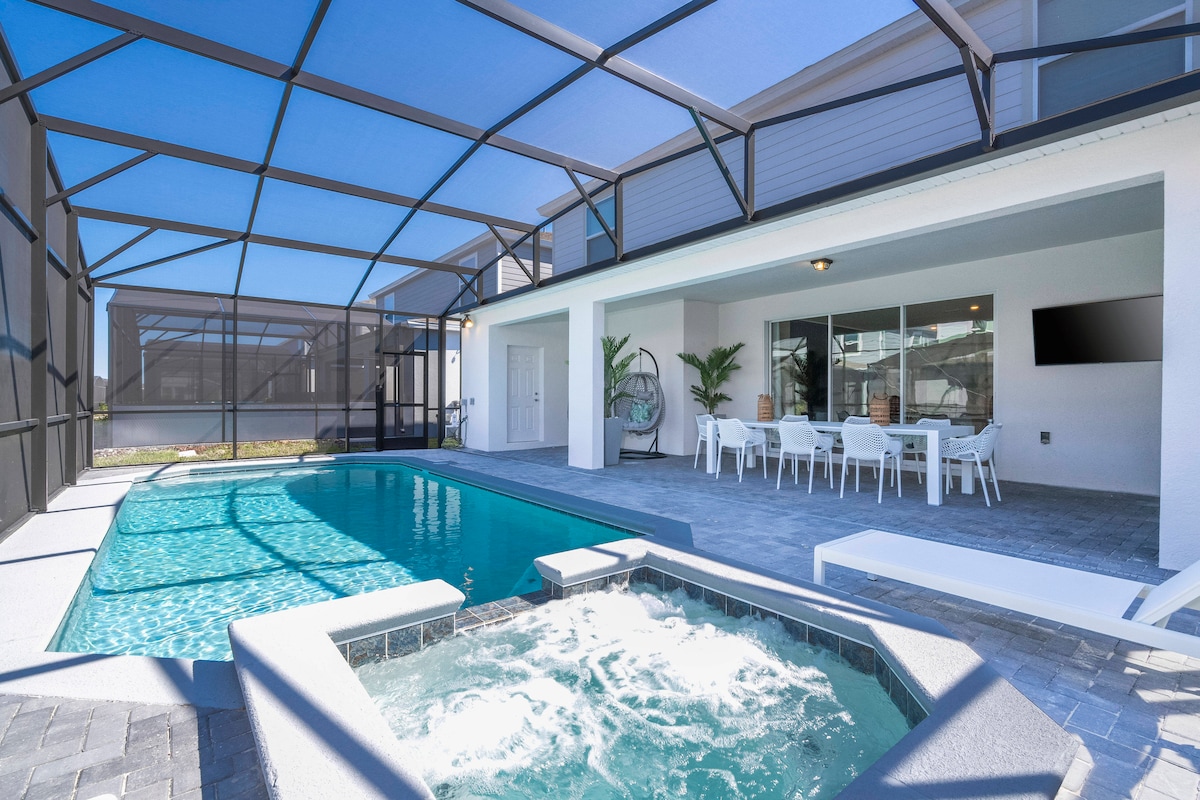 The outdoor area features a large pool and a jacuzzi, surrounded by screening to protect from insects. A dining table and chairs are visible under the covered patio, offering a shaded space for meals, while lounge chairs line the edge of the pool.