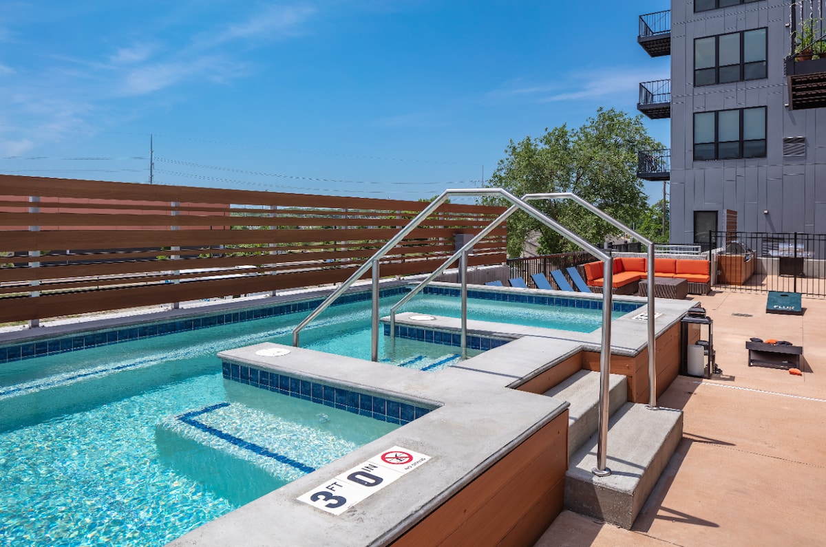A modern outdoor pool area features a landscaped wooden deck, with an in-ground pool lined by sleek steps. Loungers are positioned nearby, while vibrant orange seating provides additional relaxation options against the backdrop of the building.