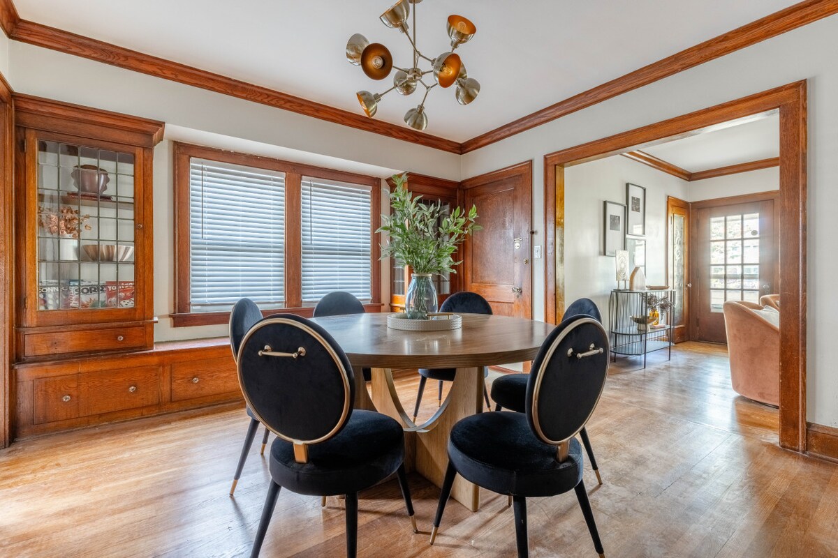 A dining area features a round wooden table surrounded by six black chairs. Natural light enters through two windows fitted with wooden blinds. A glass vase containing greenery is placed in the center of the table, complementing the warm wooden accents of the space.