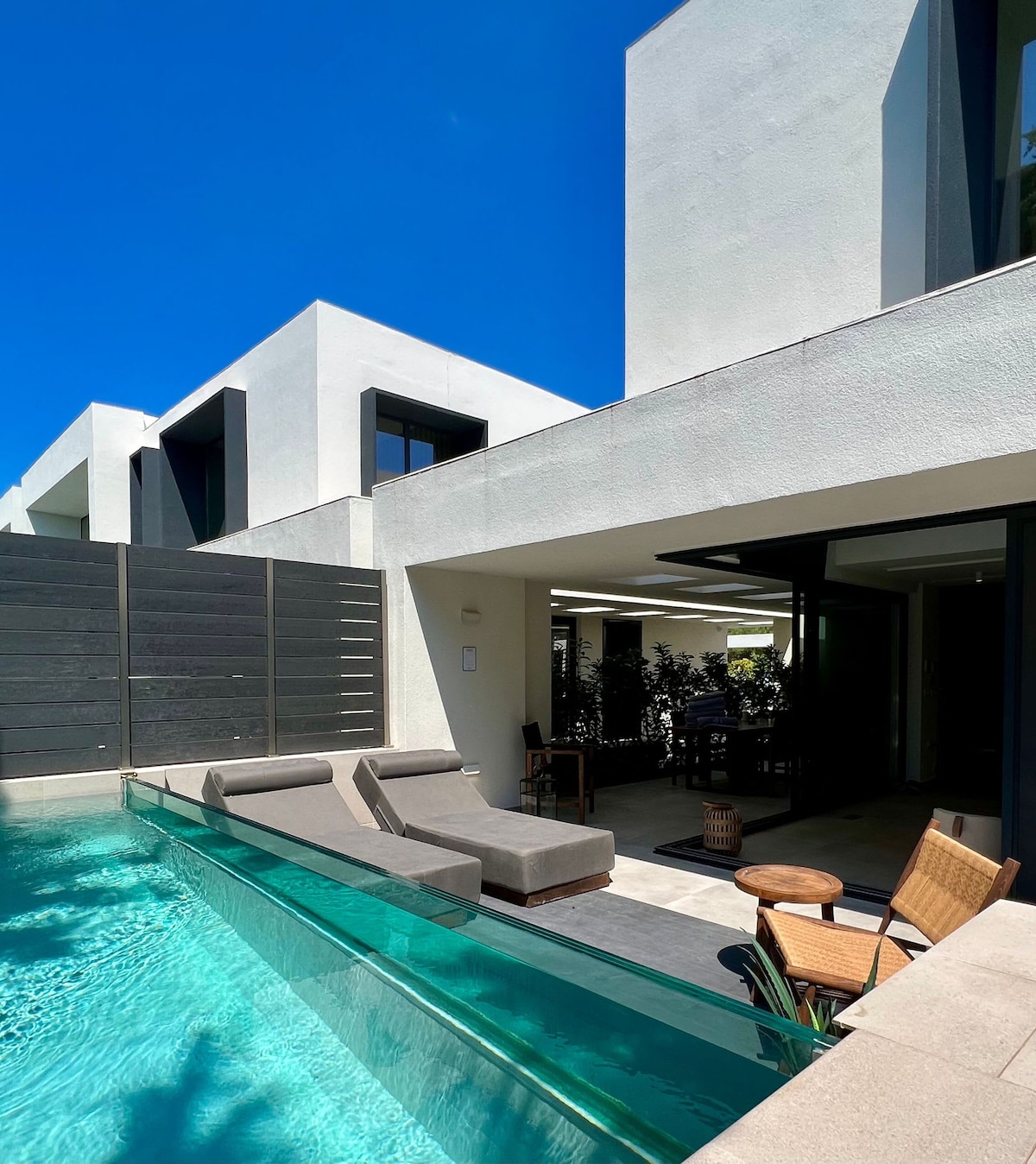 A private pool with clear water reflects the bright blue sky, bordered by modern lounge chairs. The exterior walls of the villa are finished in clean white lines, with large windows that allow natural light to fill the interior spaces.