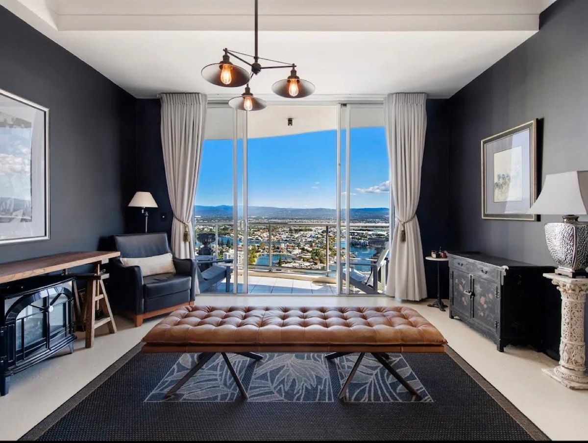A modern living area showcases a stylish leather ottoman at its center, surrounded by comfortable seating and a sleek desk. Floor-to-ceiling windows frame panoramic views of the coastline and cityscape, while soft natural light illuminates the space.