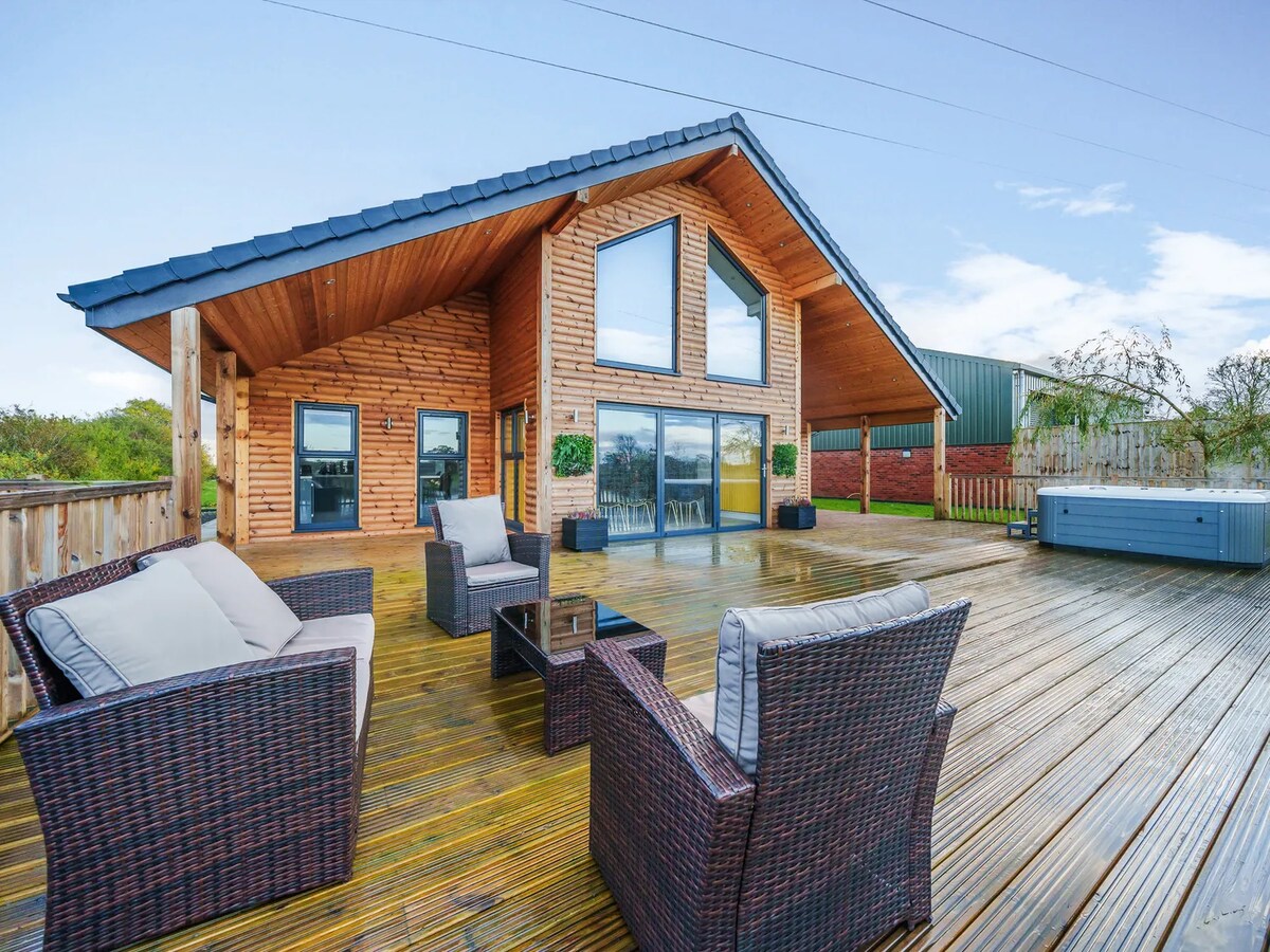 A spacious wooden deck features a combination of outdoor seating with soft cushions and a glass-topped table. The lodge's striking architectural design includes large, triangular windows that offer panoramic views, while a hot tub is positioned nearby for relaxation.