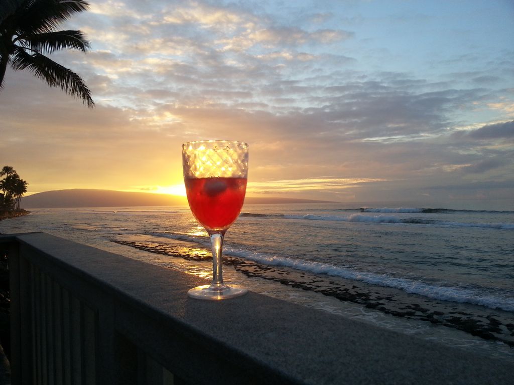 A vibrant drink is perched on a ledge, reflecting the warm hues of a sunset over the ocean. Gentle waves lap at the shoreline, while palm trees frame the scene, enhancing the serene coastal atmosphere.