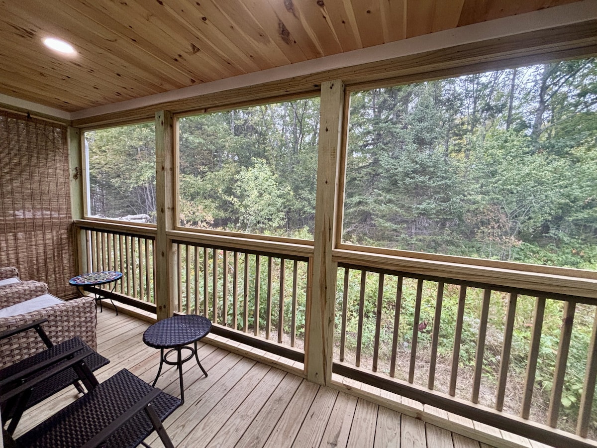 A screened porch features wooden flooring and walls, surrounded by nature with views of greenery outside. Comfortable seating is provided with two chairs and a small round table, creating an inviting space for relaxation and enjoyment of the outdoors.