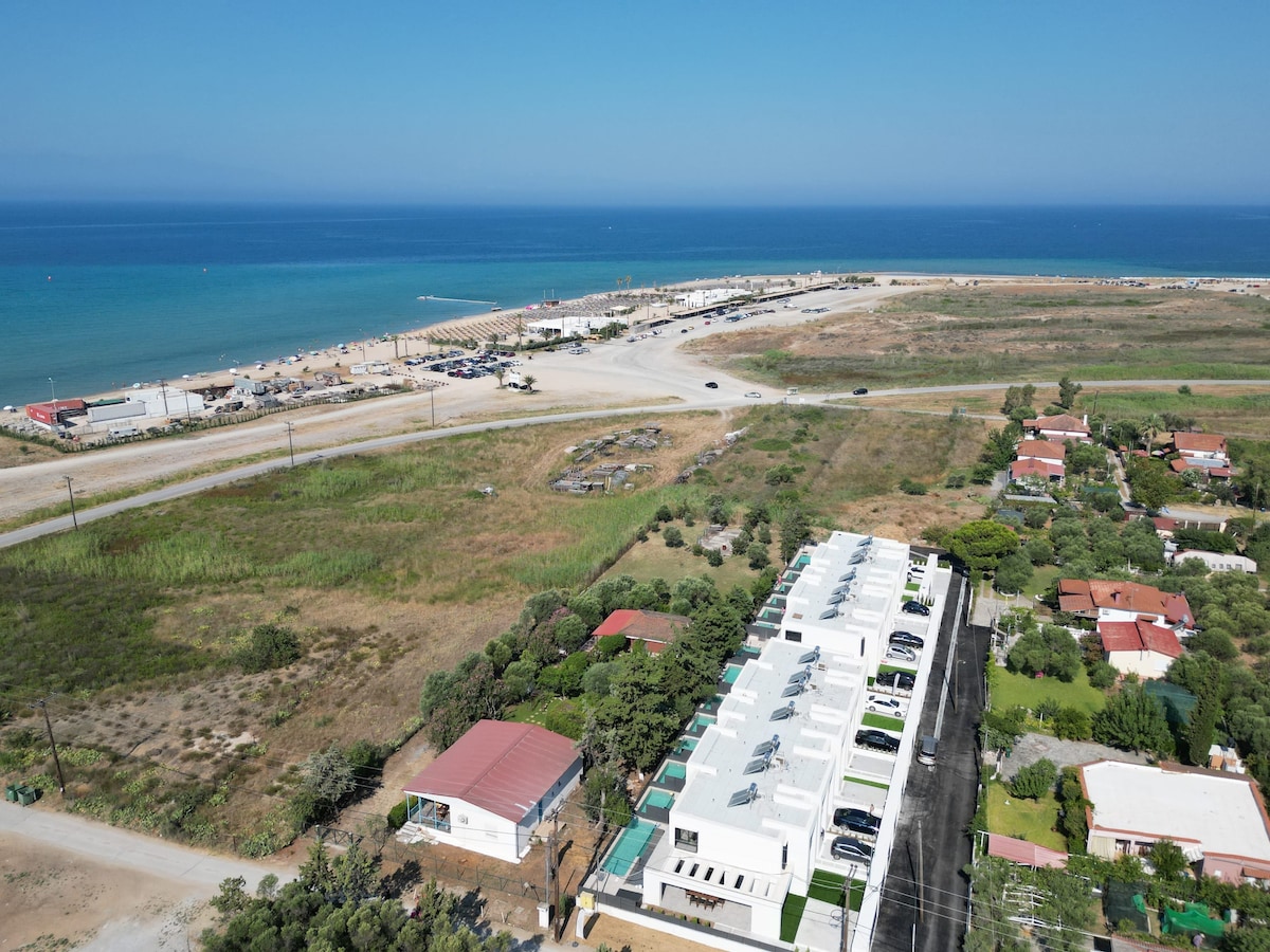 An aerial view captures the layout of the villa complex, showcasing a modern design with private pools and landscaped gardens. The coastline and clear blue waters of the beach are visible in the background, providing a contrast to the green spaces and building architecture.