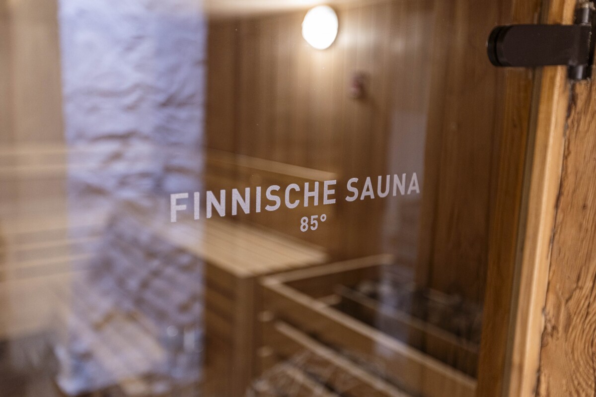 The image displays a warmly lit Finnish sauna, featuring wooden benches and paneling. The door shows the words 'Finnische Sauna 85°', indicating the sauna's temperature. The natural materials enhance the authentic sauna experience.