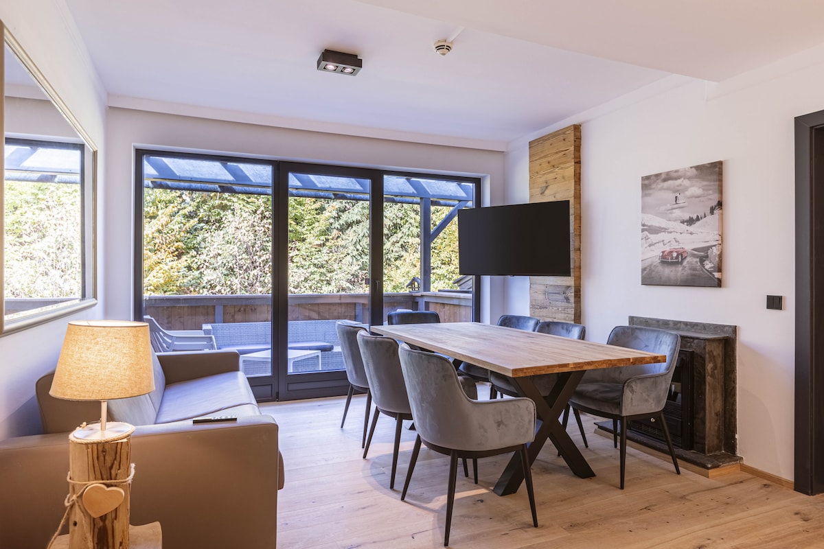 A modern living area features a wooden dining table surrounded by plush grey chairs. Large floor-to-ceiling windows provide natural light and views of the outdoors. A comfortable sofa is positioned nearby, and a wall-mounted television is visible, enhancing the suite's inviting atmosphere.