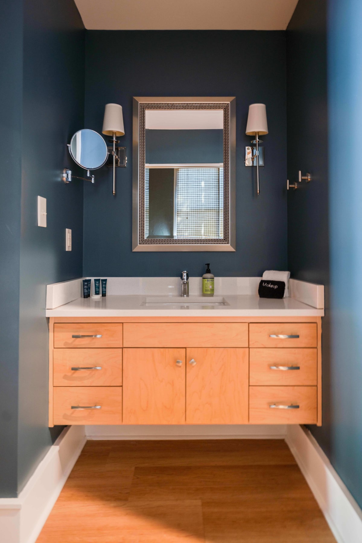 A compact bathroom space features a sleek wooden vanity with multiple drawers. The dark blue walls add depth, while a well-lit mirror is mounted above the sink. Elegant sconces frame the mirror, creating a cohesive and functional design.