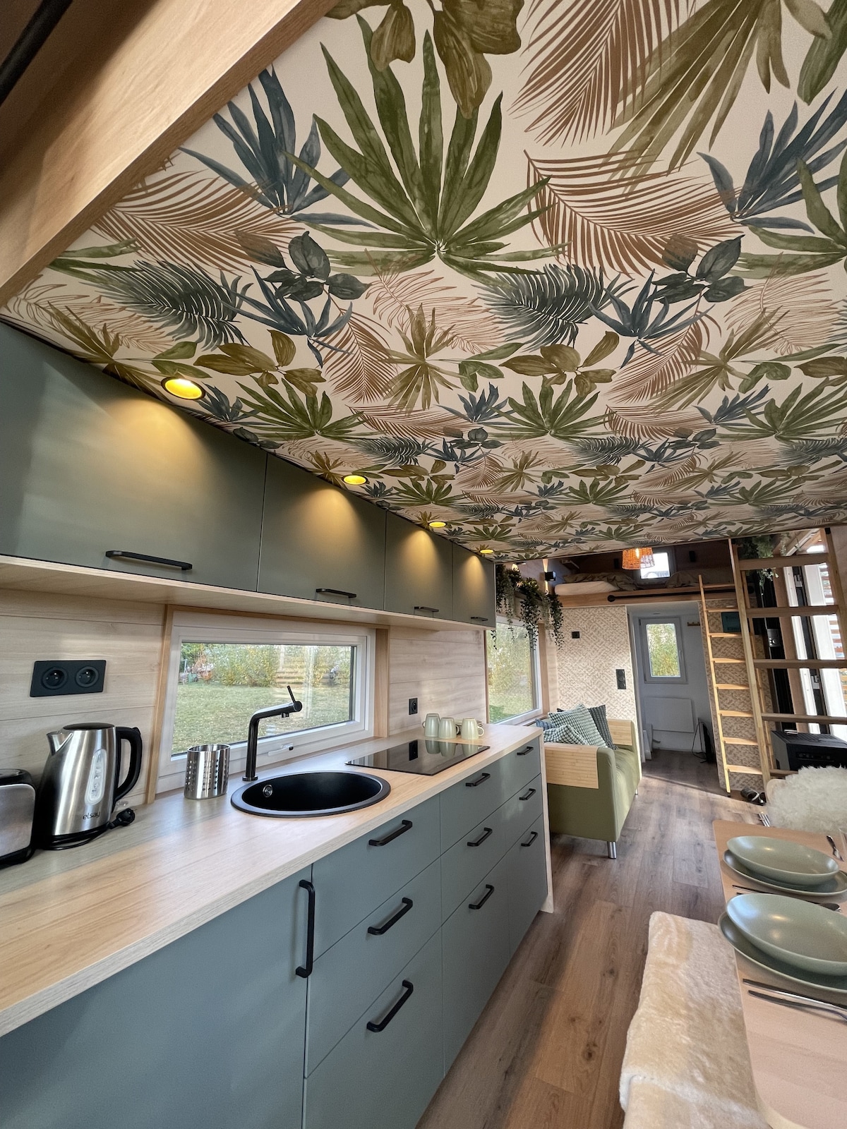 The kitchen area features stylish cabinetry with a blend of muted green and natural wood finishes. A sink and modern appliances, including a kettle and coffee maker, are presented. The ceiling is adorned with a vibrant botanical design, enhancing the overall aesthetic.
