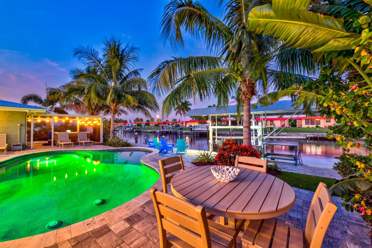 An outdoor dining area is surrounded by tropical palm trees, with a circular table and chairs placed near a heated pool. The pool features green lighting, creating a calm ambiance as it reflects the evening sky. A boat dock is visible along the tranquil water.