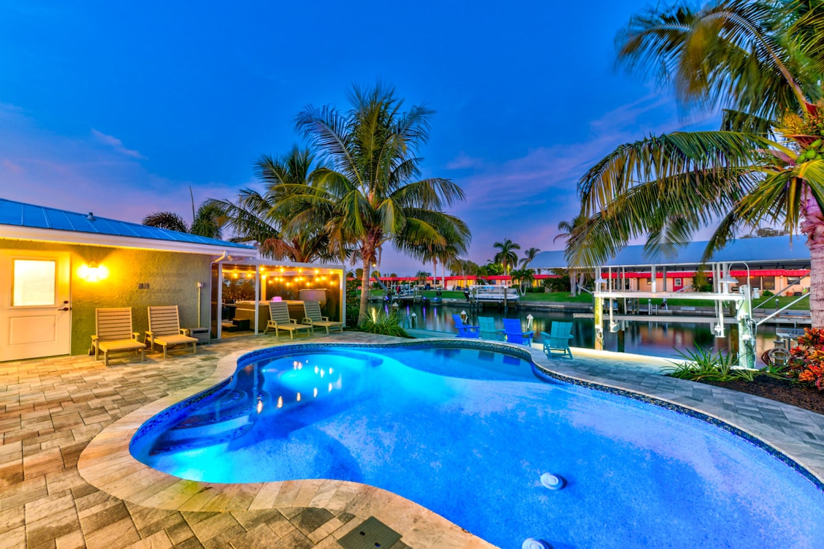 A serene backyard oasis features a heated pool with gentle lighting and a curved shape, surrounded by lounge chairs. Lush palm trees frame the space, with tranquil waters visible beyond, reflecting the evening sky's soft hues.