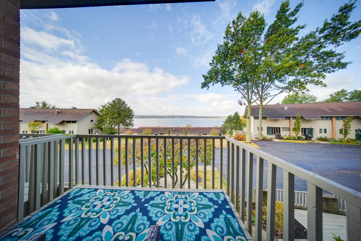 A private balcony offers expansive views over Chautauqua Lake, framed by trees and neighboring buildings. A patterned rug is placed on the balcony floor, enhancing the outdoor space. The sky is light with scattered clouds, creating a serene backdrop.